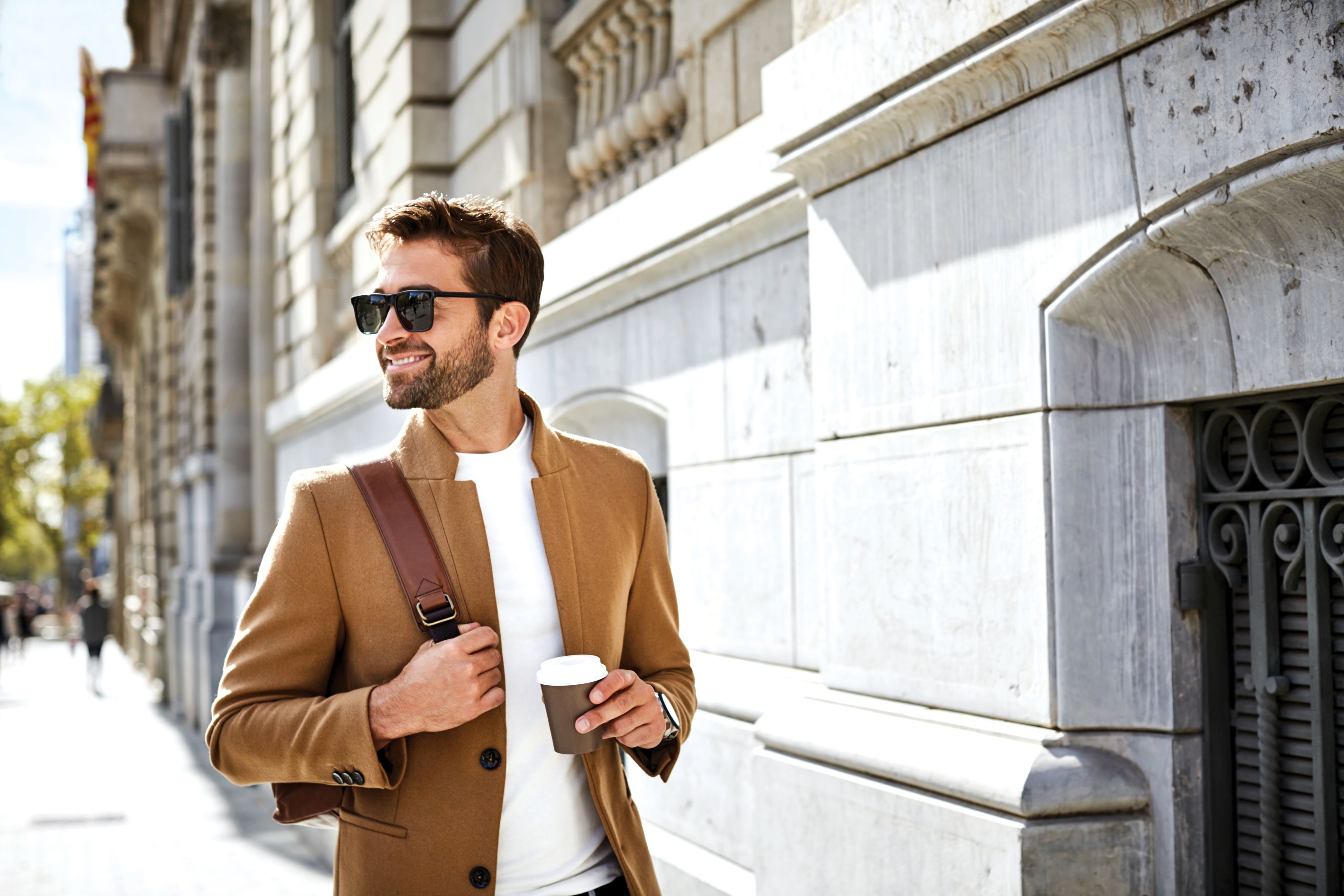 A man walking in the sun with a coffee wearing sunglasses. 