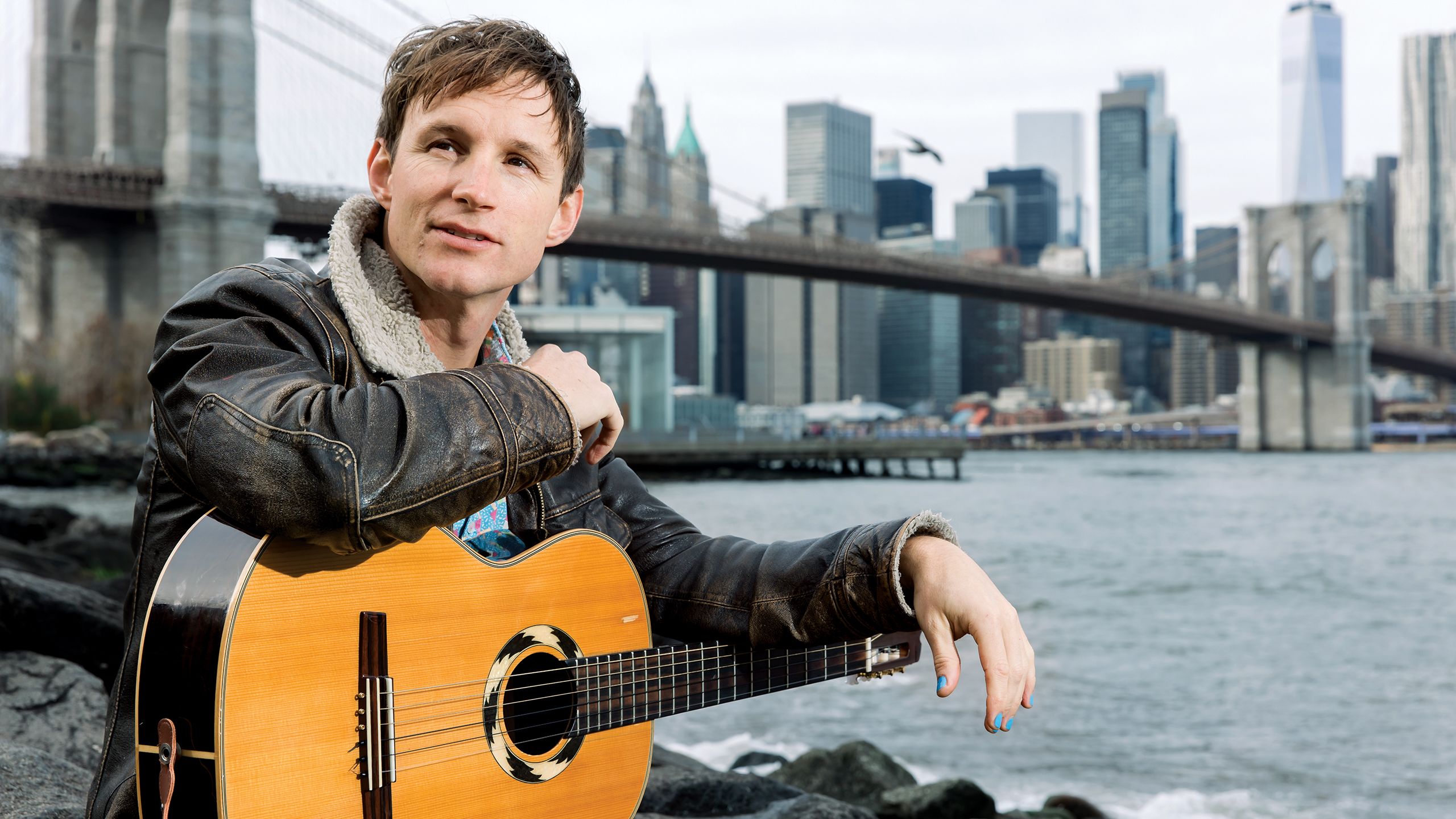 Man sitting near a bridge with a guitar.
